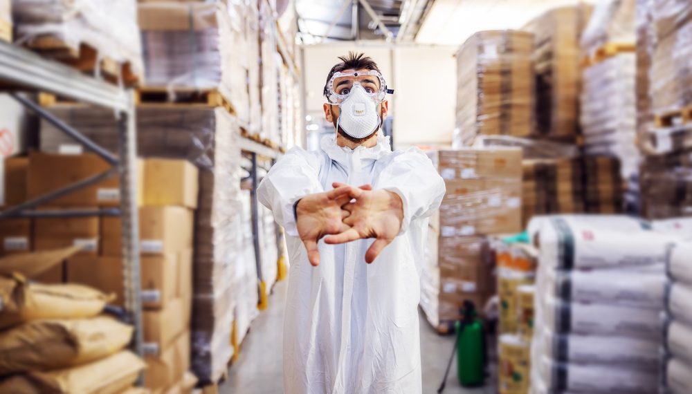 Young man in protective sterile suit and mask standing in warehouse and preparing himself to sterilize interior from corona virus/ covid-19.
