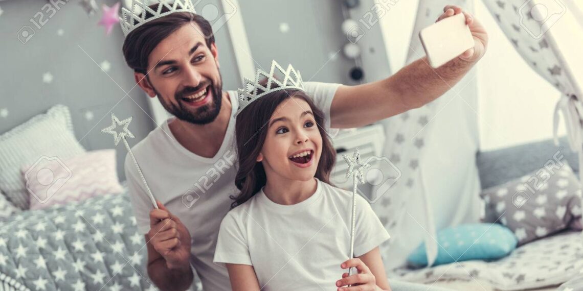 Cute little daughter and her handsome young dad in crowns are doing selfie using a smart phone and smiling while playing together in child's room