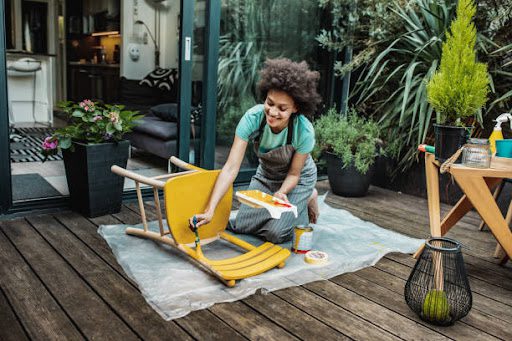 Female artist painting vintage chair in yellow color with a paintbrush in the back yard